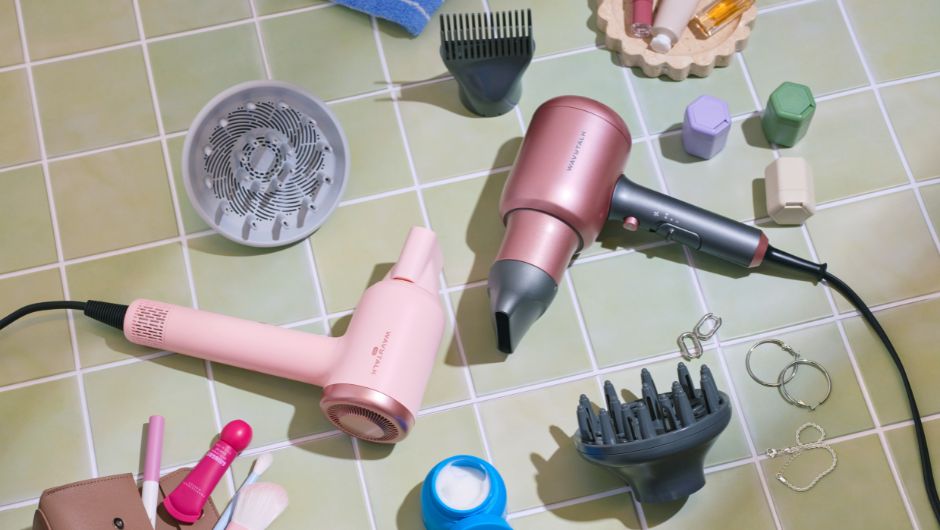 Pink hair styling tools arranged on a tiled surface, including two hair dryers, diffuser and concentrator attachments, clips, and small accessories.
