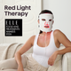 A woman wearing a white LED red light therapy face mask with red illuminated accents sits on a couch in a bright room. Large text reads 'Red Light Therapy,' and an award badge says 'ELLE Tools and Treatments Awards 2026.