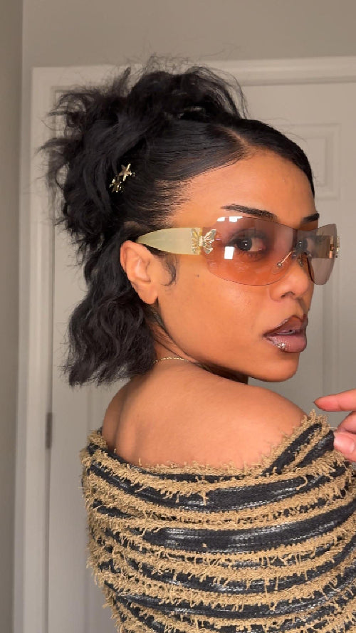 A woman indoors wearing gold sunglasses, a textured top, and a beautiful hairstyle.