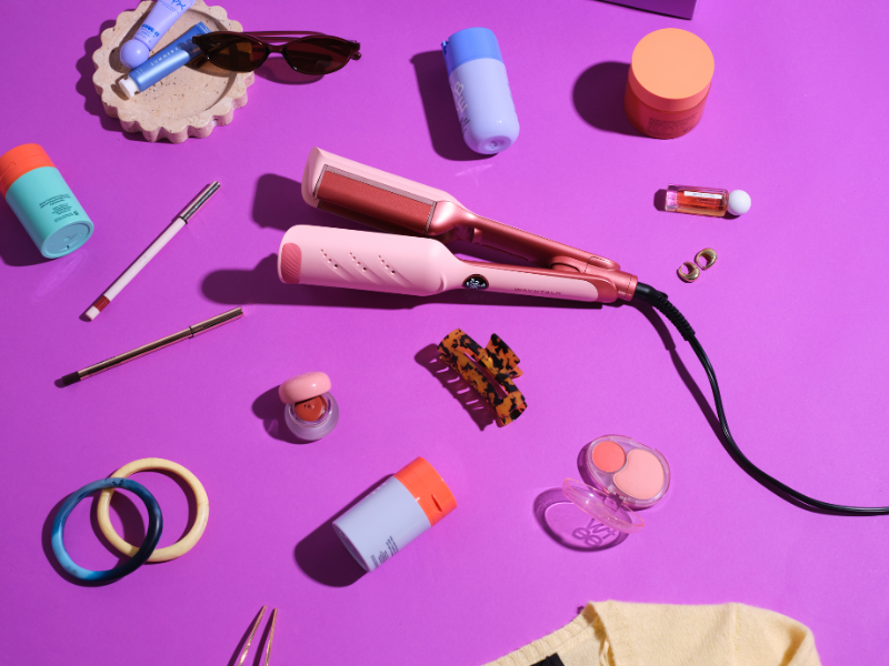 Pink hair straightener surrounded by various small items on a purple background
