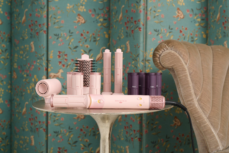 A pink hair styling tool set with multiple attachments, including a flat iron, curling barrels, and a brush, displayed on a small round table next to a beige armchair against a green floral wallpaper background.